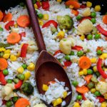 A skillet full of colorful vegetables and rice, with a wooden spoon resting in the center.