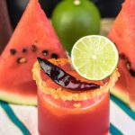 A glass of a spicy grilled watermelon margarita, garnished with a lime wedge and cayenne pepper, with a few slices of grilled watermelon in the background