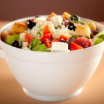 A white bowl filled with a crispy Italian salad with a variety of vegetables, feta cheese, and olives.