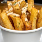 A heap of golden-brown, crispy fries arranged in a white bowl with a garlic parmesan sauce drizzled on top