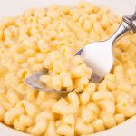 Brightly lit image of a bowl overflowing with cheesy mac and cheese with a spoon in the center