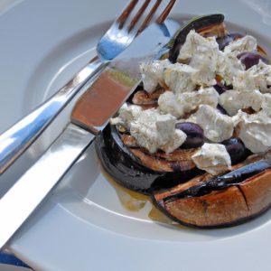 A close up of a grilled eggplant with feta and olives on a plate with a knife and fork