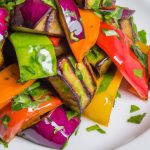 A close-up image of a grilled eggplant salad with a variety of colorful vegetables and herbs on a white plate.