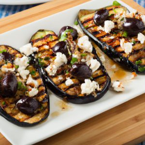 A plate with grilled eggplant topped with feta cheese and olives on a cutting board.