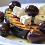 A close-up of a grilled eggplant dish with feta cheese and olives on a white plate.