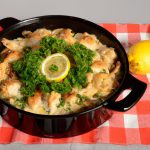 A large skillet of Comforting French Chicken Casserole with a lid on top, garnished with fresh parsley and wedges of lemon, on a black and white checkered tablecloth.