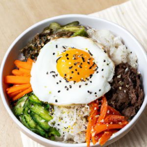 A bowl of bibimbap with colorful vegetables, rice, and a sunny-side-up egg, garnished with sesame seeds, and served on a light wood table.