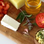 A wooden cutting board with freshly cut ingredients for a frittata, including tomatoes, feta cheese, herbs and olive oil.