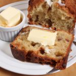 A golden-brown loaf of apple fritter bread cut into thick slices and served with a dollop of butter.