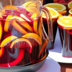 A close-up of a pitcher of red sangria with a lemon and orange slices in it, and several glasses of red sangria with a variety of fruit slices and herbs in them.