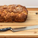 A loaf of golden brown apple fritter bread on a wooden board with a knife beside it.
