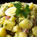 Image of a large bowl of French Potato Salad, with potatoes, pickles, eggs, and herbs. It is topped with fresh parsley.