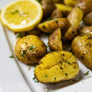 A plate of roasted lemon-herb potatoes with a bright yellow lemon sliced in half, fresh herbs, and a sprinkle of salt.