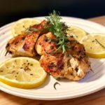 A plate of grilled Italian chicken, with a sprig of rosemary, garlic, and lemon slices on top.