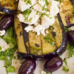 A close up of grilled eggplant with feta and olives on a plate with a side of fresh greens.