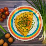 A plate with a perfectly cooked French omelette in the center, sprinkled with fresh chives, surrounded by a colorful array of fresh vegetables.