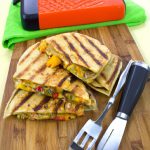 Grilled veggie quesadillas on a cutting board with a pair of tongs