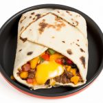 A cast iron skillet with a half-wrapped breakfast burrito in the center, with warm egg, cheese, and peppers on a white background.