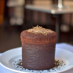 A tall, perfectly risen French chocolate soufflé sitting atop a white plate, with a few chocolate shavings sprinkled on top.