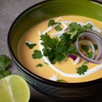 A bowl of creamy coconut curry soup with cilantro, onion and lime slices on top.