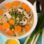 A bowl of Chinese Egg Drop Soup with a spoon in the center surrounded by scallions, carrots, and mushrooms.