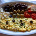 A large plate of Mediterranean-style breakfast appetizers including an omelette, tomatoes, olives, feta cheese, and herbs