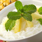 A bowl of Chinese coconut rice pudding decorated with a few slices of pineapple, a sprinkle of toasted coconut flakes and a sprig of mint.