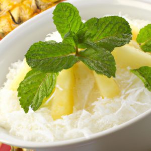 A bowl of Chinese coconut rice pudding decorated with a few slices of pineapple, a sprinkle of toasted coconut flakes and a sprig of mint.