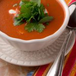 A bowl of Mexican Roasted Red Pepper Soup with a spoon and a sprig of parsley for garnish.