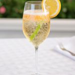 A glass of Italian Prosecco Spritz on a white tablecloth with a lemon wedge and fresh herbs in the background.