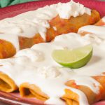 A plate of classic Mexican enchiladas with a side of guacamole, sour cream, and limes.