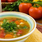 A bowl of Mediterranean Soup with carrots, celery, onions, tomatoes, and herbs in the background.