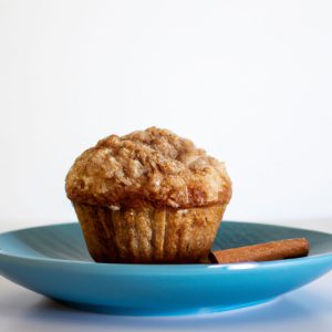 A warm, freshly-baked apple-cinnamon muffin on a light blue plate with a white background and a sprinkle of cinnamon on top.