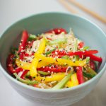 A bowl of colorful Chinese sesame noodle salad topped with sesame seeds, green onions, and red pepper flakes.