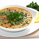 A large white bowl filled with steaming Mediterranean Lentil Soup. The bowl is garnished with fresh parsley, wedges of lemon and a sprinkling of chili flakes.