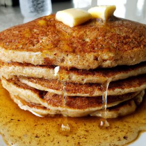 A stack of fluffy, golden-brown oatmeal pancakes with a pat of butter, a drizzle of syrup, and a sprinkle of cinnamon on top.