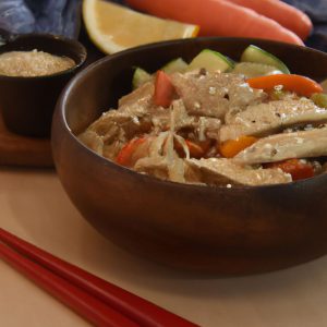 A bowl of Chinese Sesame Chicken Noodle Bowl with a wooden spoon and a plate of vegetables to the side.