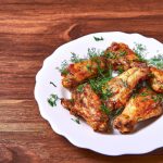 A plate of golden brown oven-baked chicken wings with herbs on a white plate against a wooden background.