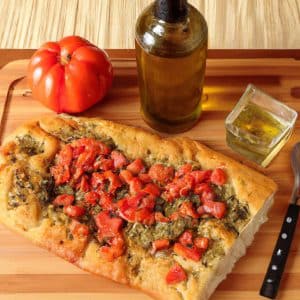 A freshly baked Italian Tomato Basil Focaccia served on a cutting board with a wooden spoon and an olive oil bottle.